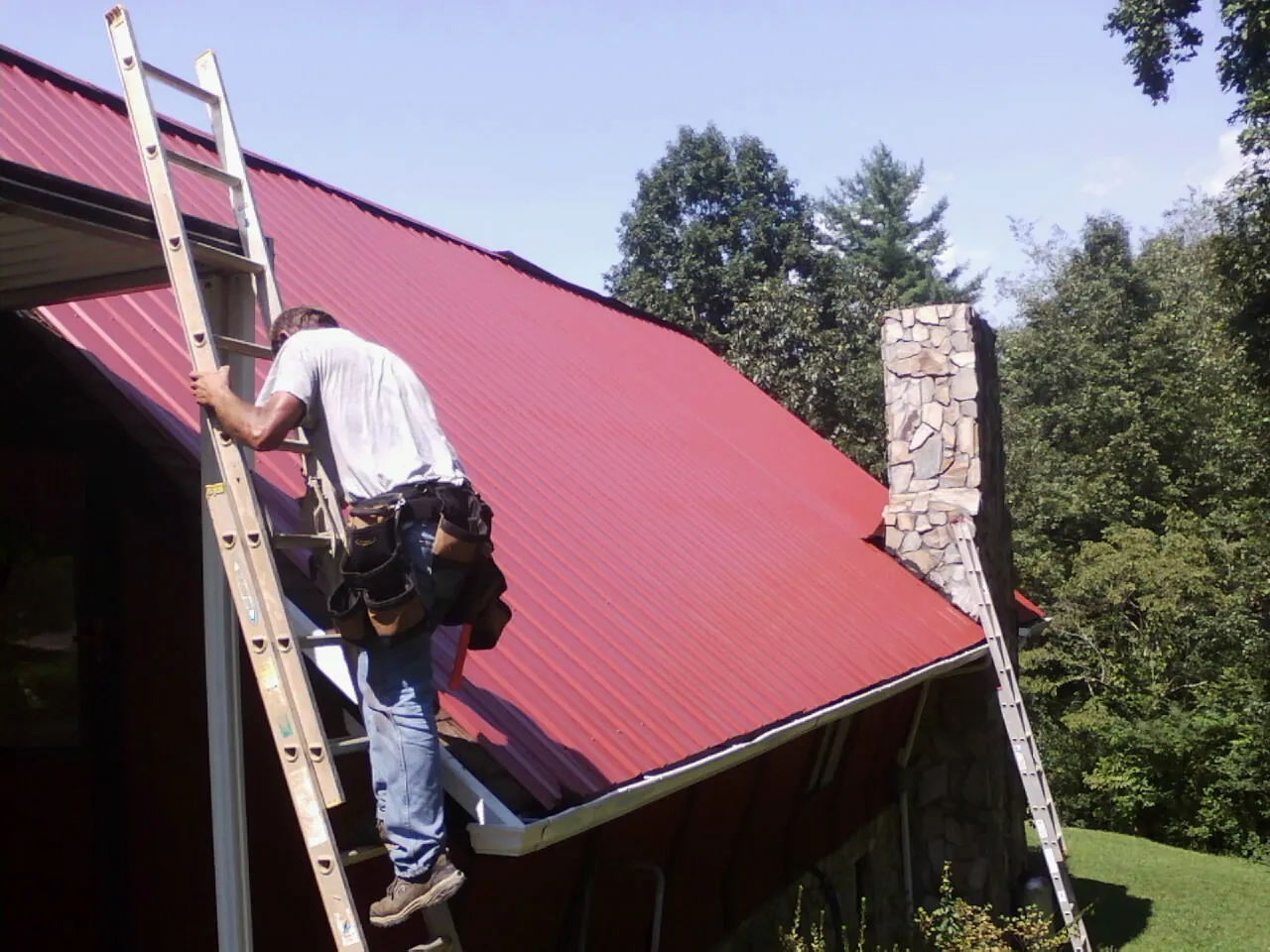 Roofer on a ladder beside a red metal roof and stone chimney — Soffit & Fascia Repair by Roofing Brothers in Hiwassee